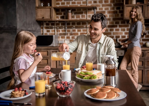 Familia feliz desayunando en la cocina. Papá e hija están sentados a la mesa mientras mamá prepara panqueques.