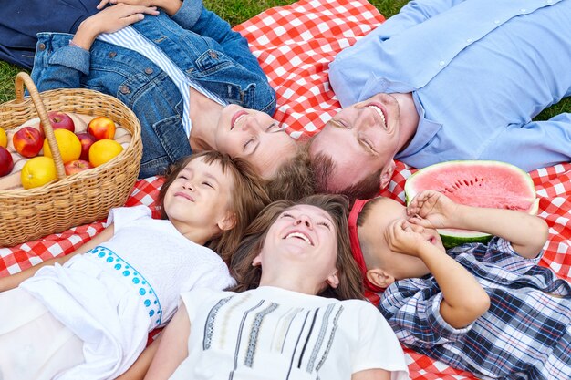 Familia feliz descansando en un picnic. Disfrutando y acostado sobre cuadros escoceses en la pradera. Los adultos y los niños miran al cielo.