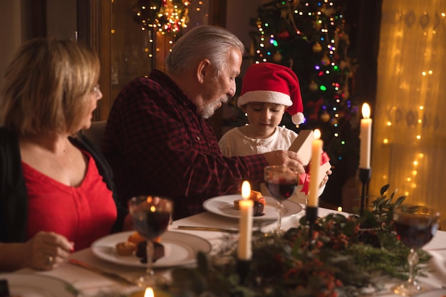 Família feliz desfrutando do jantar de Natal em casa perto da árvore de Natal. Avô sênior dando um presente para seu pequeno neto com chapéu de Papai Noel. É hora de abrir os presentes.
