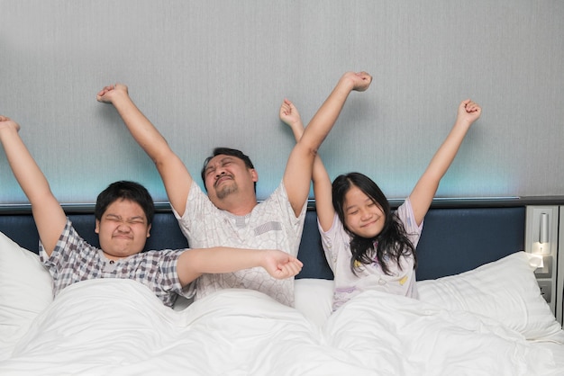 La familia feliz se despierta por la mañana estirando la mano hacia arriba mientras está sentado en la cama en el fondo del dormitorio. Padre caucásico con su hijo y su hija se ejercitan en la cama.