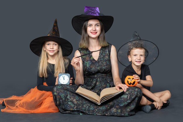 Una familia feliz con disfraces espeluznantes de brujas y magos sosteniendo un gran libro con predicciones, un reloj negro y una varita mágica en un fondo negro Víspera de Halloween