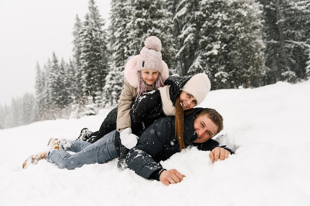 Familia feliz divertirse en el bosque de invierno. Madre, padre e hija yacen en el suelo y juegan con la nieve. Concepto de familia. Disfrutando pasar tiempo juntos