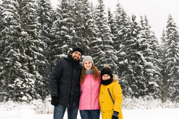 Familia feliz divertirse en el bosque de invierno y mirando a cámara. Madre, padre e hijo jugando con nieve. Concepto de Navidad familiar. Disfrutando pasar tiempo juntos