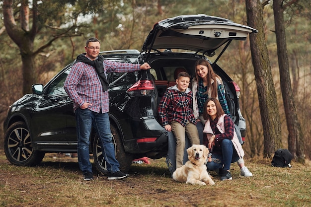 La familia feliz se divierte con su perro cerca de un auto moderno al aire libre en el bosque