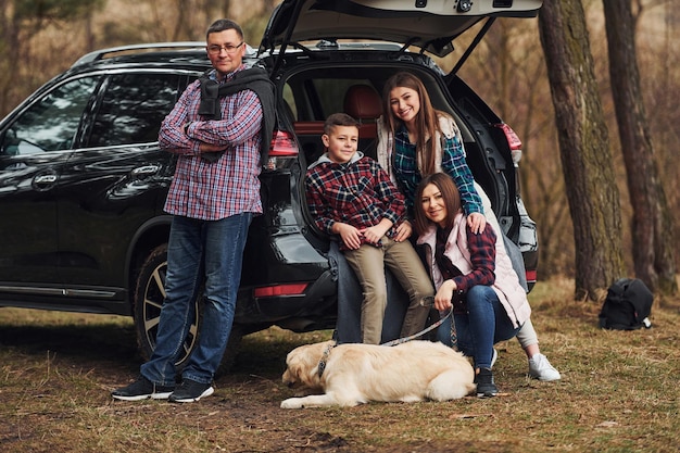 La familia feliz se divierte con su perro cerca de un auto moderno al aire libre en el bosque