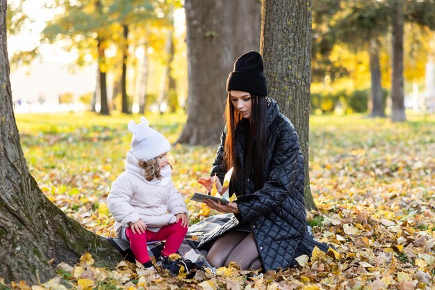 Familia feliz divirtiéndose en el parque de otoño Una madre está leyendo un libro con su linda hija mientras está sentada en el césped del parque