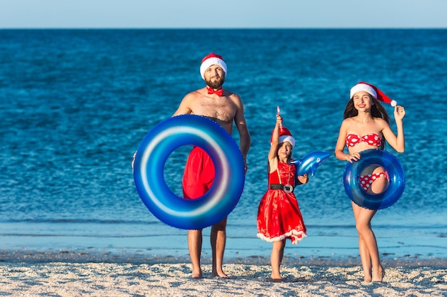 Família feliz e alegre, vestida com chapéu de Papai Noel e com círculos infláveis, festeja o Natal de verão na praia do mar.