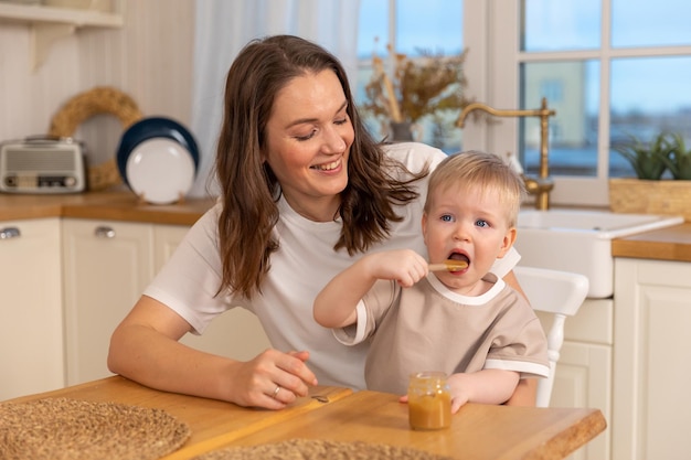 Família feliz em casa mãe alimentando bebê na cozinha garotinho com cara engraçada bagunçada come fo