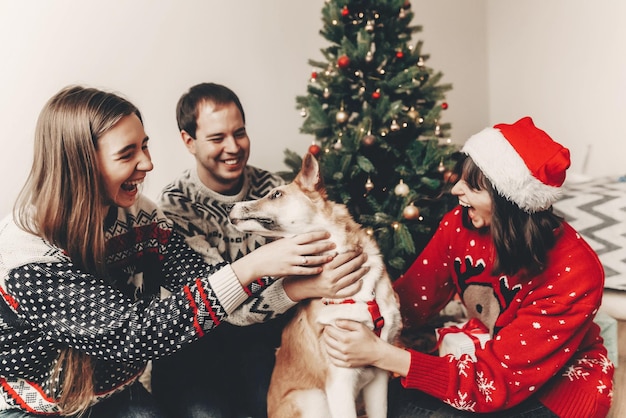Foto família feliz em suéteres elegantes e cachorro fofo se divertindo na árvore de natal com luzes momentos emocionais atmosféricos feliz natal e feliz ano novo espaço de conceito para texto