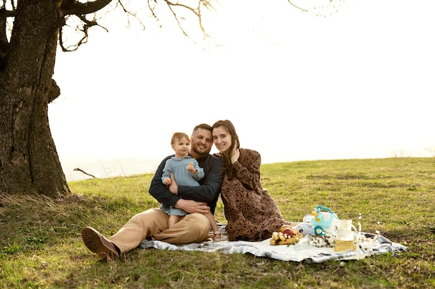 Família feliz em um piquenique um pai com uma esposa grávida e uma filha pequena estão na natureza no parque ao pôr do sol família jovem está descansando no gramado comida e bebidas para um piquenique casal positivo