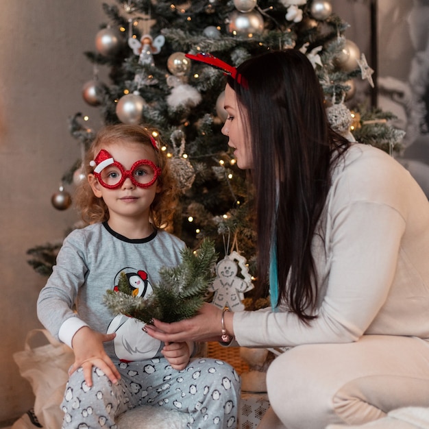 Família feliz engraçada, mãe e filha em pijama da moda sentar-se perto de árvores de Natal com luzes. Férias de inverno em casa