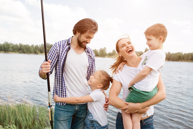 Família feliz está de pé juntos. Mulher está rindo alto e segurando o filho nas mãos dela. Pequena menina está abraçando o pai dela. Cara está segurando uma vara de peixe na mão direita.