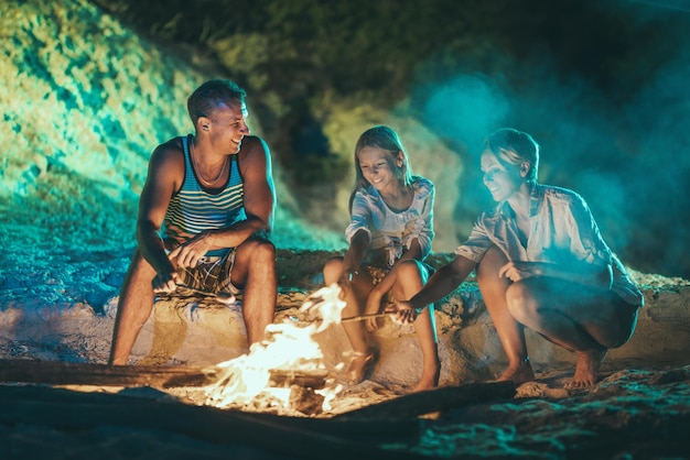 Una familia feliz está sentada en la orilla del mar junto al fuego por la noche. Están friendo salchichas en palo.