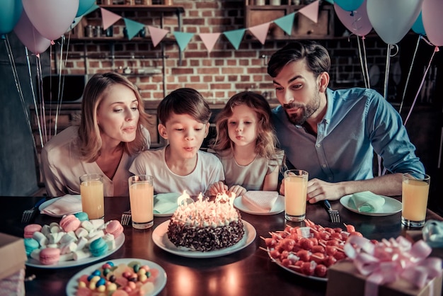 La familia feliz está soplando velas en el pastel mientras está sentado a la mesa en la cocina decorada durante la celebración del cumpleaños