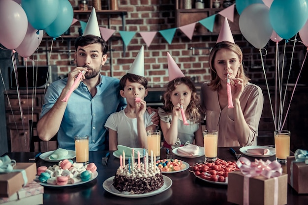 Família feliz está soprando em chifres de festa na câmera enquanto está sentado à mesa na cozinha decorada durante a festa de aniversário