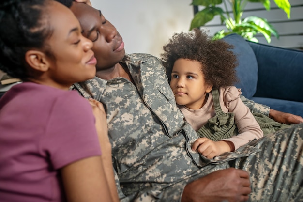 Familia feliz. Feliz militar de piel oscura y una mujer bonita con los ojos cerrados y mirando a la pequeña hija linda en casa en el sofá