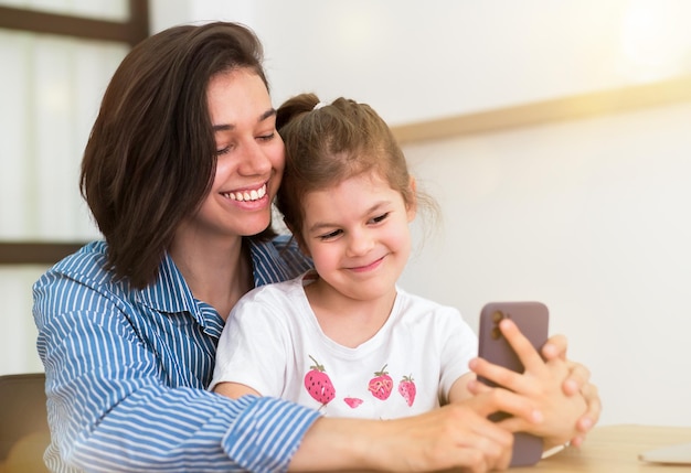 Foto família feliz, filha fofa, com a mãe rindo usando o smartphone assistindo um vídeo engraçado no aplicativo de mídia social tirando uma selfie olhando para a tela do celular em casa