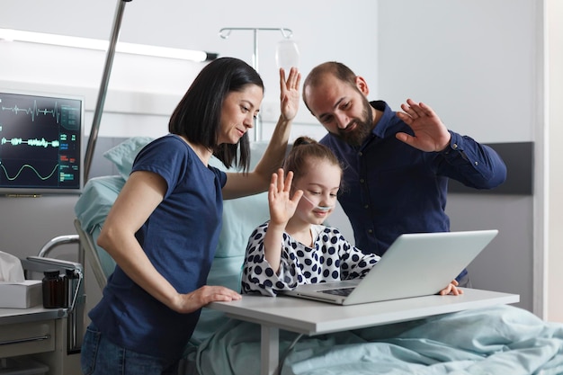 Familia feliz hablando con parientes a través de la tecnología de videollamadas mientras está en la habitación del hospital. Niña enferma y padres seguros de sí mismos saludando en una teleconferencia en línea mientras están sentados en la sala de pacientes.