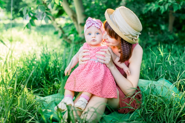 Una familia feliz haciendo un picnic en el jardín verde en un soleado día de primavera: una hermosa madre sonriente sentada sobre la hierba verde y su pequeña hija riendo en sus piernas