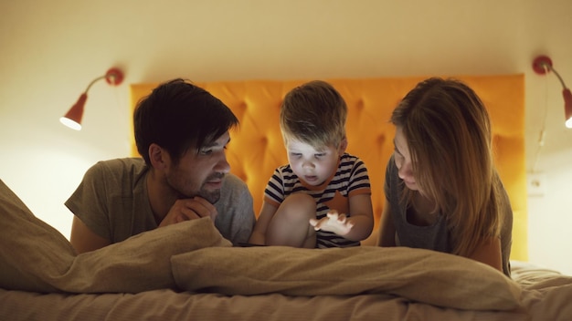 Familia feliz con un hijo pequeño aprendiendo a jugar a la computadora de la tableta acostado en la cama en casa por la noche