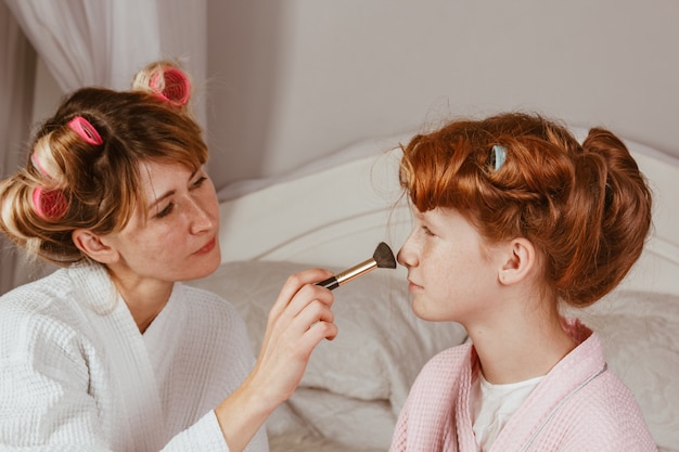 Família feliz. jovem mãe linda faz maquiagem de sua filha linda na cama no quarto. a filha ruiva com rolinhos na cabeça está rindo.