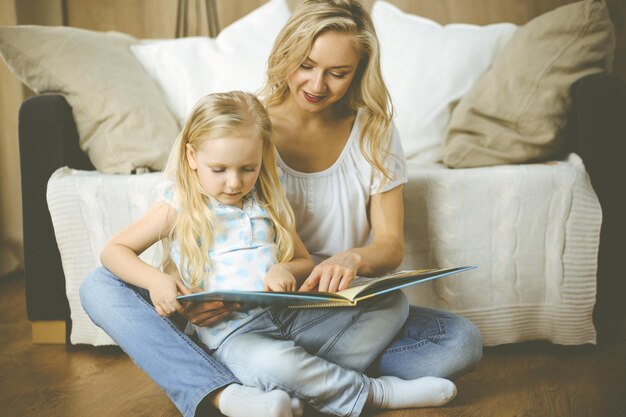 Familia feliz. Joven madre rubia leyendo un libro a su linda hija mientras se sienta en el suelo de madera. Concepto de maternidad.