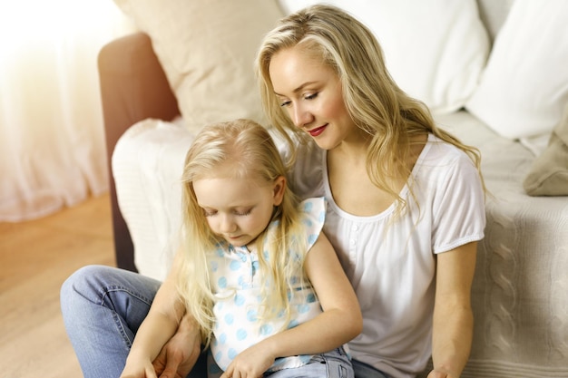 Familia feliz. Joven madre rubia leyendo un libro a su linda hija mientras se sienta en el suelo de madera en una habitación soleada. Concepto de maternidad.