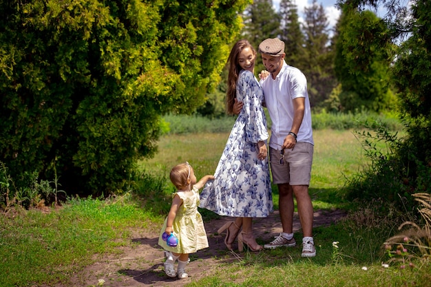 Familia feliz juega con la pequeña hija en el parque verde. Niña sostiene el vestido de su madre al aire libre, horario de verano