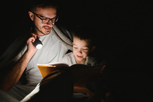 Família feliz lendo história de ninar sob o cobertor à noite. Pai e filho passam tempo juntos. Dia dos Pais.