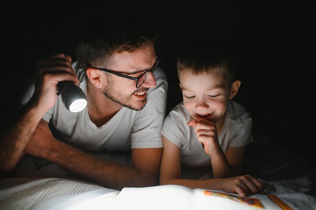 Familia feliz leyendo un cuento antes de dormir bajo una manta por la noche Padre e hijo pasan tiempo juntos el Día del Padre