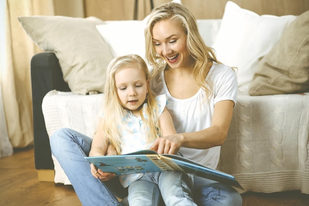 Família feliz. Loira jovem mãe lendo um livro para sua filha bonita enquanto está sentado no chão de madeira. Conceito de maternidade.