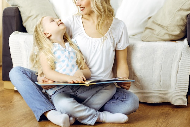 Família feliz. Loira jovem mãe lendo um livro para sua linda filha enquanto está sentado no chão de madeira. conceito de maternidade