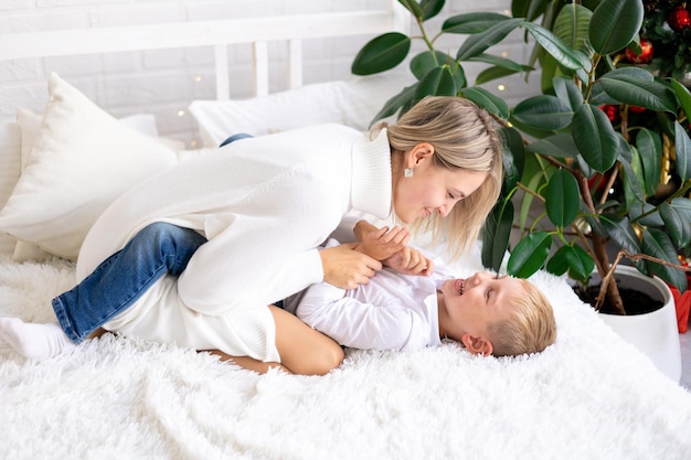 Familia feliz madre e hijo con suéteres blancos y jeans en la cama en un dormitorio luminoso en casa abrazándose y sonriendo