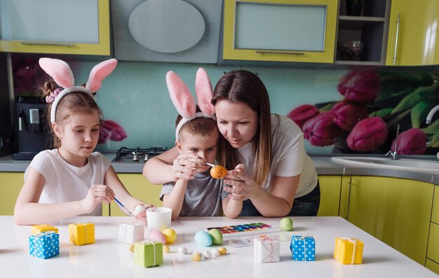 Familia feliz: madre, hija e hijo con orejas de conejo se preparan para las vacaciones, coloreando huevos en la acogedora cocina de la casa. preparativos para la fiesta de Pascua