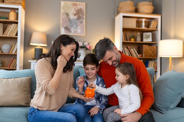 Familia feliz madre y padre alegres con hijos sonriendo y poniendo monedas en la alcancía mientras están sentados en el sofá en casa