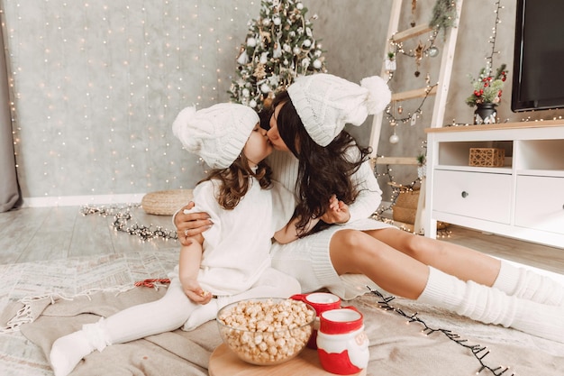 Família feliz, mãe e menina com chapéus de malha brancos sentados perto de uma árvore de Natal com um presente de Natal