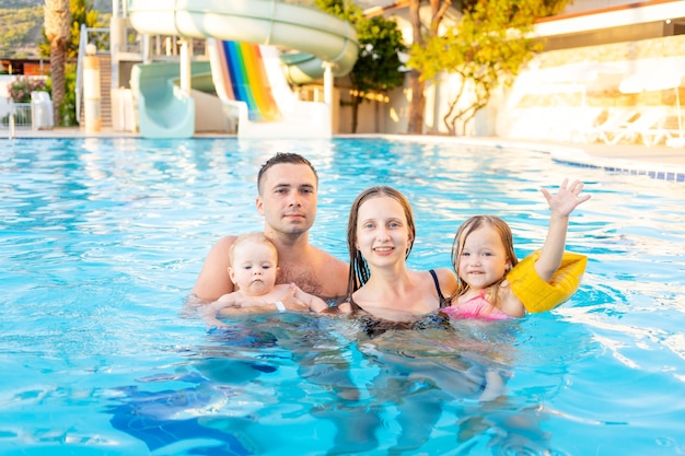 Família feliz, mãe, pai e dois filhos estão nadando na piscina com toboáguas e se divertindo nas férias, sorrindo e rindo