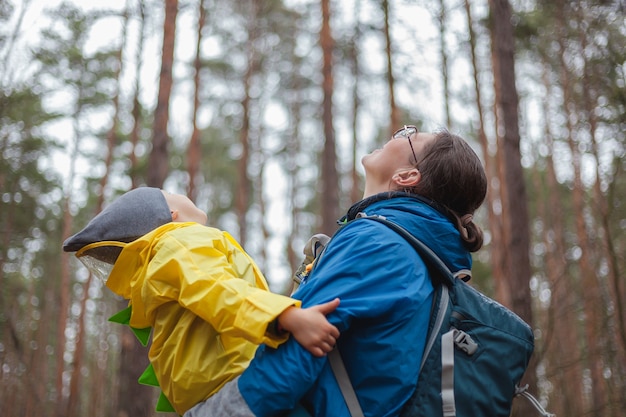 Familia feliz Mamá y niño caminan en el bosque después de la lluvia en impermeables juntos, abrazan y miran al cielo