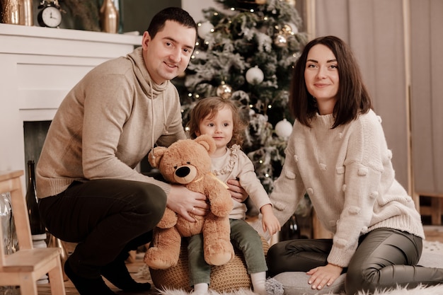 Foto familia feliz, mamá, papá e hija pequeña con oso de peluche cerca del árbol de navidad y chimenea en casa.