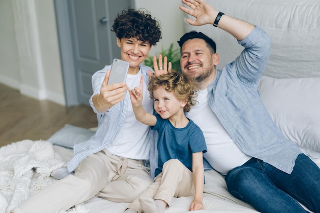 Familia feliz mamá papá e hijo tomando selfie en la cama agitando las manos sonriendo