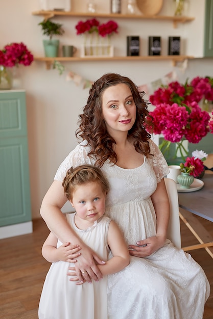 Familia feliz, mujer embarazada con pequeña hija en la cocina con flores de peonías. Relación entre padres e hijos. Maternidad, embarazo, concepto de felicidad.