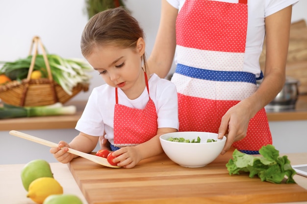 Família feliz na cozinha Mãe e filha filha cozinhando saboroso café da manhã de salada fresca Pequeno ajudante cortando e misturando tomates e hortaliças