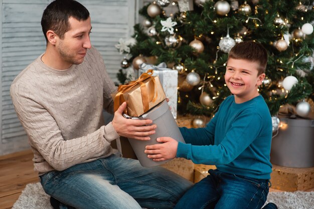 Una familia feliz con un niño celebra un año nuevo o Navidad. Padre e hijo se sientan en el suelo cerca del árbol de Navidad en el hogar e intercambian regalos