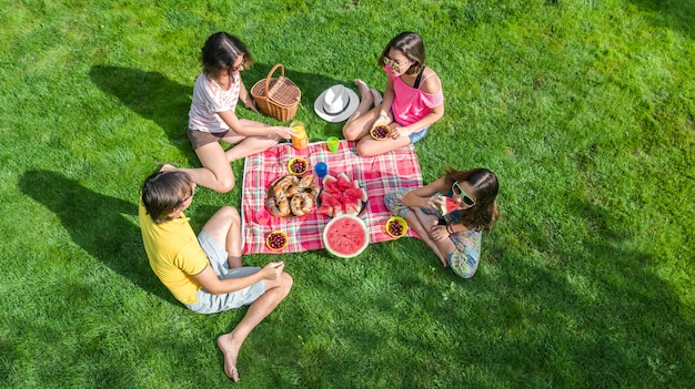 Familia feliz con niños haciendo un picnic en el parque, padres con niños sentados en el césped del jardín y comiendo comidas saludables al aire libre, vista de aviones no tripulados desde arriba, vacaciones familiares y concepto de fin de semana