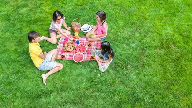 Familia feliz con niños haciendo un picnic en el parque, padres con niños sentados en el césped del jardín y comiendo comidas saludables al aire libre