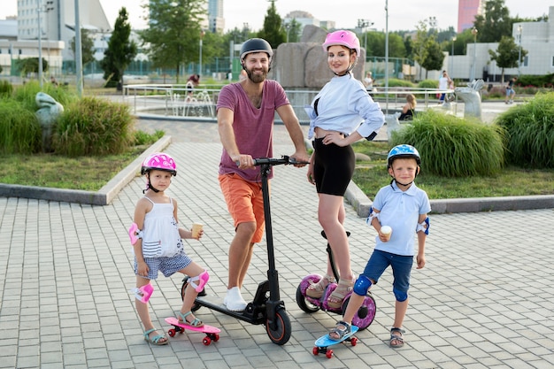 Familia feliz con niños en Segways, scooter eléctrico y patinetas en el parque en verano, niños comiendo helado.