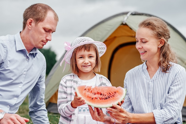 Família feliz no piquenique no camping. mãe, pai e filha comendo melancia perto de uma barraca no prado ou parque