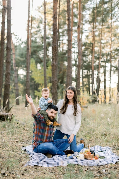 Familia feliz, padre guapo, madre atractiva y alegre hijo pequeño, divirtiéndose juntos en el bosque de otoño, sentados en una manta con frutas