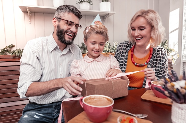 Familia feliz. Padres positivos felices sentados alrededor de su hija mientras le dan un regalo