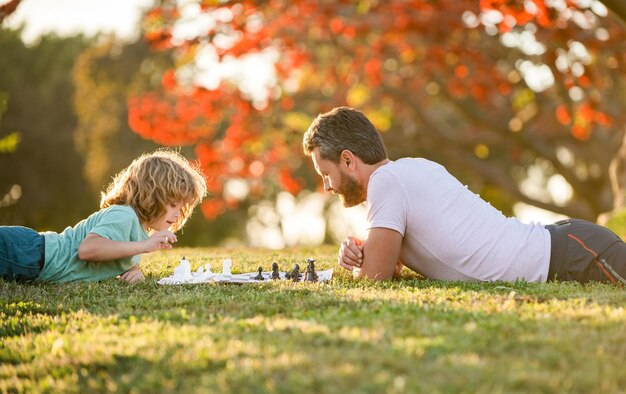 Familia feliz de papá e hijo jugando al ajedrez en la hierba verde en el parque al aire libre, erudito